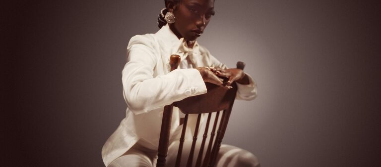 Flo Milli sits in a chair wearing all white. She's looking at the camera.
